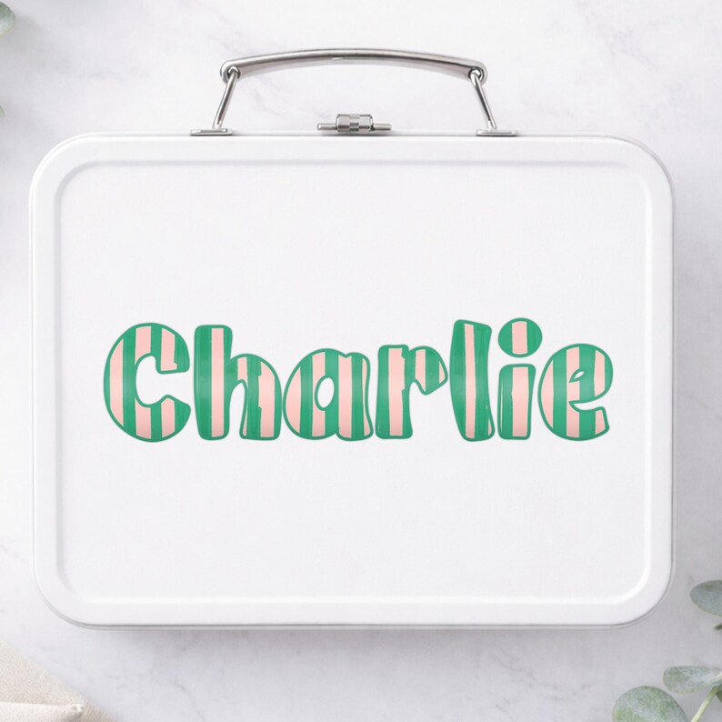 Op de afbeelding: Witte metalen lunchbox met een zilveren handvat. De naam "Charlie" is in grote, retro-stijl letters op de voorkant gedrukt. De letters zijn groen met roze verticale strepen, wat een speels en gepersonaliseerd ontwerp cre&euml;ert.