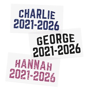 Puede incluir: Tres pegatinas rectangulares con nombres y fechas. La pegatina superior dice "CHARLIE 2021-2026" en purpurina azul. La pegatina del medio dice "GEORGE 2021-2026" en negro. La pegatina inferior dice "HANNAH 2021-2026" en purpurina rosa.