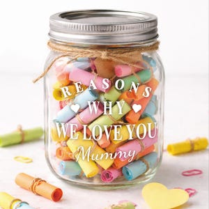 May include: A clear glass jar with a silver lid, filled with colorful rolled-up paper notes. The jar is decorated with twine and features the text "REASONS WHY WE LOVE YOU Mummy" in white lettering. Several additional rolled notes are scattered around the jar.