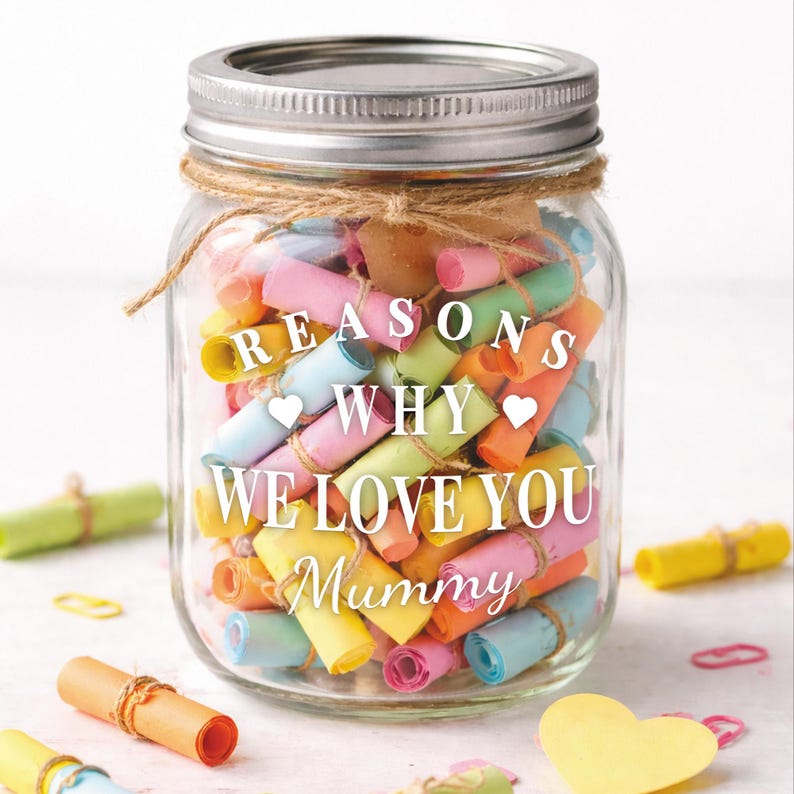 May include: A clear glass jar with a silver lid, filled with colorful rolled-up paper notes. The jar is decorated with twine and features the text "REASONS WHY WE LOVE YOU Mummy" in white lettering. Several additional rolled notes are scattered around the jar.