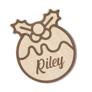 Pu&ograve; includere: Un ornamento natalizio in legno marrone a forma di budino con un ramo di agrifoglio in cima. L'ornamento ha il nome "Riley" scritto su di esso.