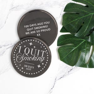 May include: Two black round coasters with silver trim. One coaster reads "I QUIT Smoking 100 DAYS AGO" with star accents. The other coaster reads "100 DAYS AGO YOU QUIT SMOKING! WE ARE SO PROUD XX".
