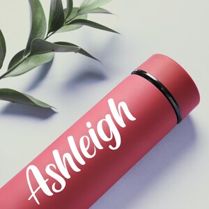 May include: A red insulated water bottle with the name "Ashleigh" in white script. The bottle has a black band near the top and is set against a light background with green leaves.