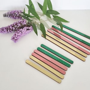 May include: A set of wooden craft sticks with glitter in gold, pink and green colours. The sticks are arranged in a staggered pattern.