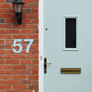 May include: Light blue metal house numbers '57' fixed to a red brick wall beside a light blue door. A black lantern is mounted above the numbers. The door features a small window and a brass letterbox.