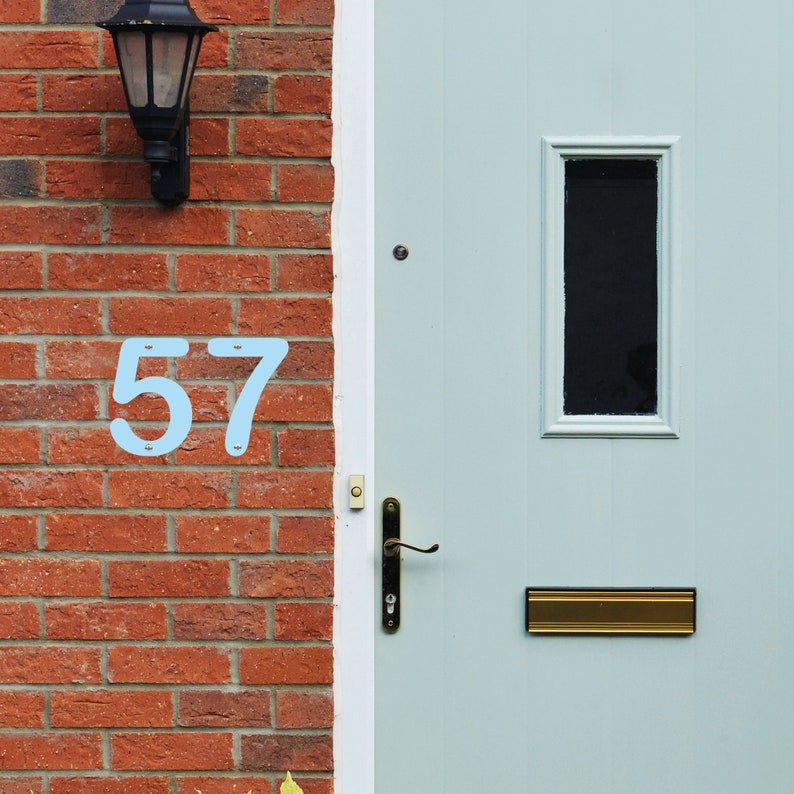 May include: Light blue metal house numbers '57' fixed to a red brick wall beside a light blue door. A black lantern is mounted above the numbers. The door features a small window and a brass letterbox.