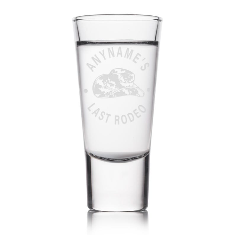 K&ouml;nnte beinhalten: Klares Schnapsglas mit dem wei&szlig;en Schriftzug "ANYNAME'S LAST RODEO". Das Design beinhaltet eine Cowboyhut-Illustration. Das Glas ist zylindrisch mit einem dicken Boden, perfekt f&uuml;r einen feierlichen Drink.