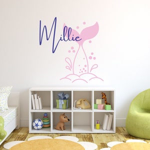 May include: Pink mermaid tail wall decal with the name Millie in blue script. The decal is on a white wall with a white bookshelf and a green armchair in the background.