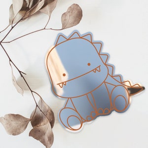 May include: A light blue mirror with a cute cartoon dinosaur design. The dinosaur is outlined in brown and has a small smile.