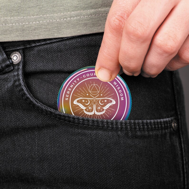May include: A round, iridescent coin with the words "SERENITY-COURAGE-WISDOM" encircling a central design. The design features a moth with moon and star details, and geometric shapes. The coin is being placed into a black denim pocket.