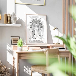 Puede incluir: Una ilustraci&oacute;n en blanco y negro de la carta del tarot La Emperatriz. La Emperatriz se representa como una mujer con una corona de flores y un vestido fluido. La carta est&aacute; enmarcada y colgada en una pared sobre un escritorio de madera.