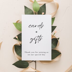 May include: A white sign with the words "cards + gifts" in elegant black script, surrounded by green leaves, small pink flowers, and a gold ribbon. The sign also includes the text "Thank you for joining us on our special day."
