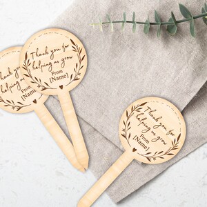 May include: Wooden stick with a round, brown, engraved tag that says "Thank you for helping us grow. From [Name]" with a small heart at the bottom. The tag has a decorative wreath around the text.