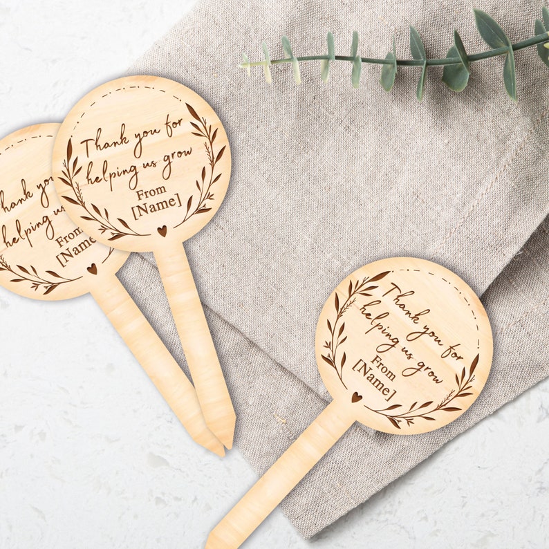 May include: Wooden stick with a round, brown, engraved tag that says "Thank you for helping us grow. From [Name]" with a small heart at the bottom. The tag has a decorative wreath around the text.
