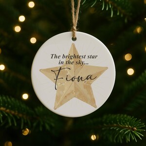 May include: A round, white Christmas ornament with a gold star design and the name "Fiona" in cursive. The text "The brightest star in the sky..." is above the star. The ornament hangs from a natural twine string.
