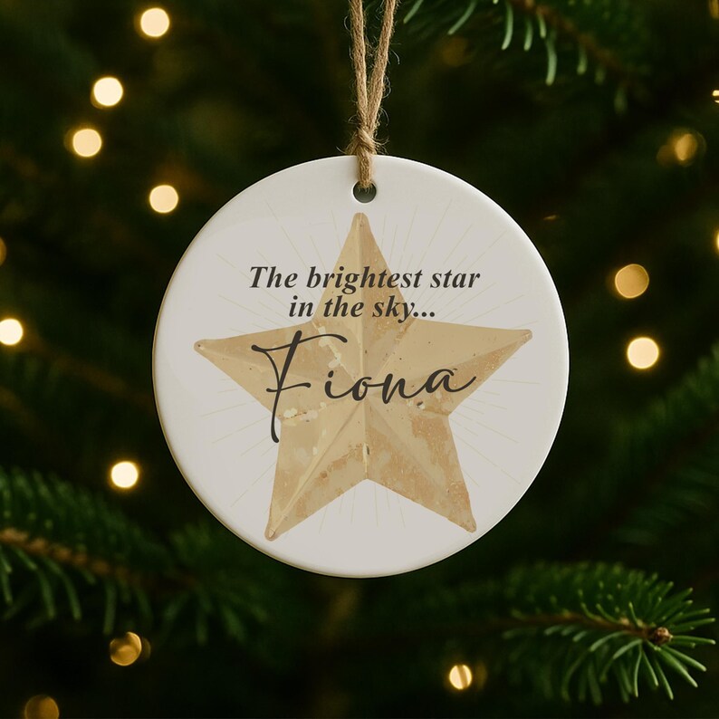 May include: A round, white Christmas ornament with a gold star design and the name "Fiona" in cursive. The text "The brightest star in the sky..." is above the star. The ornament hangs from a natural twine string.