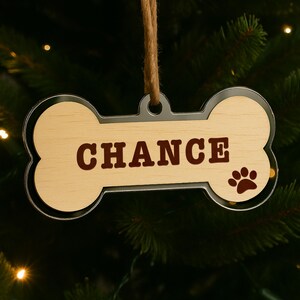 Peut inclure: Ornement en forme d'os pour chien avec le nom "CHANCE" en lettres marron. L'ornement a un centre en grain de bois clair, un bord argent&eacute; et une empreinte de patte. Une ficelle marron est attach&eacute;e pour l'accrocher.