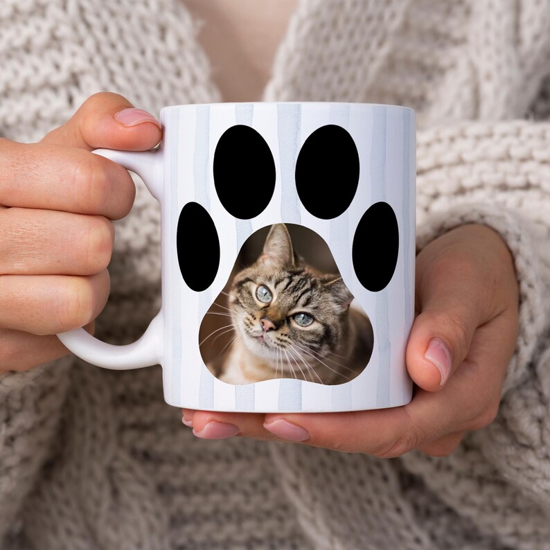 May include: White ceramic mug featuring a cat paw print design with a tabby cat portrait in the center. The mug has a white handle and is decorated with vertical light blue stripes. A charming gift for cat lovers.