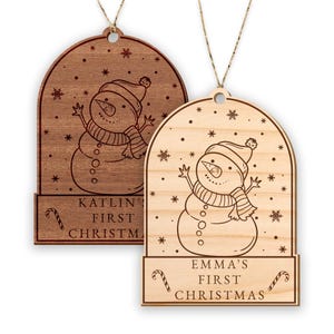 May include: Two wooden Christmas decorations featuring a snowman design. The ornaments are personalised with the text "KATLIN'S FIRST CHRISTMAS" and "EMMA'S FIRST CHRISTMAS".