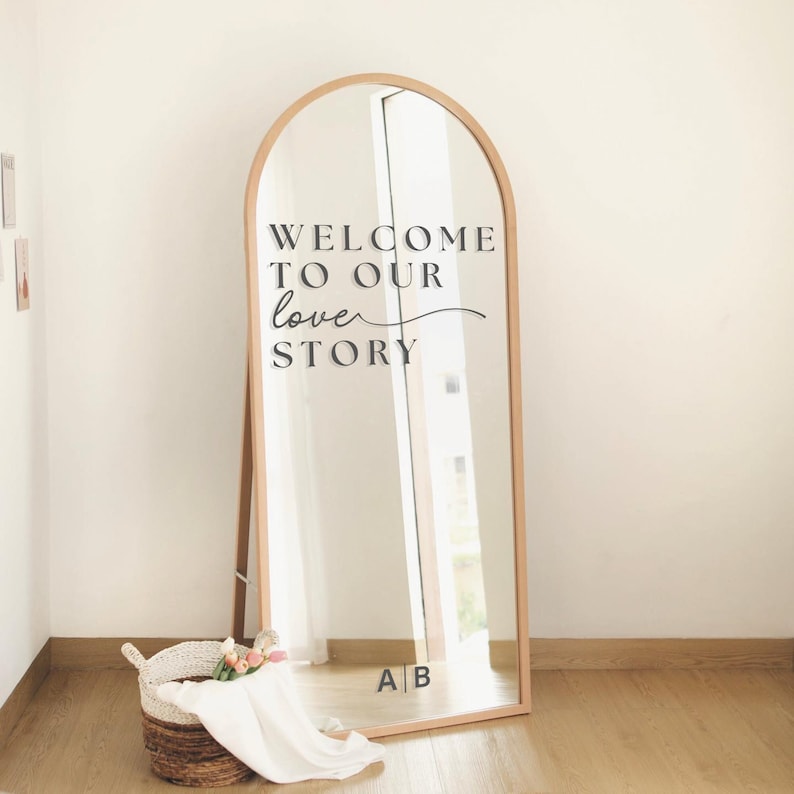 May include: A large, arched-top mirror with a wooden frame. The mirror has the text "WELCOME TO OUR love STORY" in black script. A woven basket with flowers and fabric sits in front of the mirror. The initials "A B" are at the bottom.