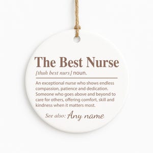 May include: A round, white ceramic ornament with the text "The Best Nurse" in brown. The ornament includes a definition of a nurse and a space for a name. A natural twine loop is at the top for hanging.