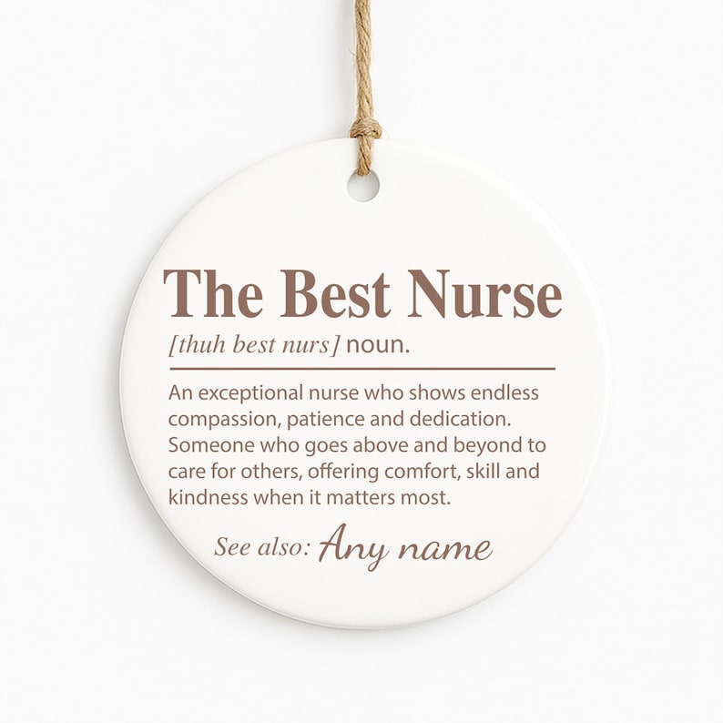 May include: A round, white ceramic ornament with the text "The Best Nurse" in brown. The ornament includes a definition of a nurse and a space for a name. A natural twine loop is at the top for hanging.