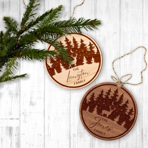 Op de afbeelding: Twee ronde houten kerstversieringen met een hert-silhouet in een bos sc&egrave;ne. De bovenste versiering zegt "The Langton Family" en de onderste versiering zegt "The Jones Family".