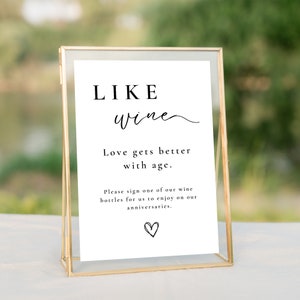 May include: A gold-framed sign with the text "LIKE wine Love gets better with age. Please sign one of our wine bottles for us to enjoy on our anniversaries." and a heart symbol.