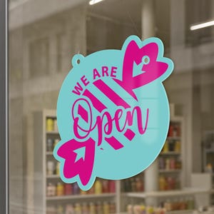 May include: A round, light blue hanging sign with the words "WE ARE Open" in pink script. The sign has a decorative pink arrow design. The sign is hanging in a window.