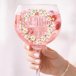 Puede incluir: Una copa grande y transparente llena de una bebida rosa y hielo. La copa est&aacute; decorada con una guirnalda de margaritas y las palabras "Best Mum Ever!". La copa es sostenida por una persona.