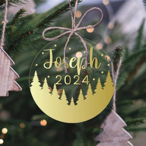 Może przedstawiać: Okrągła, przezroczysta ozdoba choinkowa z wzorem z złotej folii. Ozdoba przedstawia złotą sylwetkę lasu z sosnami i tekst "Joseph 2024".