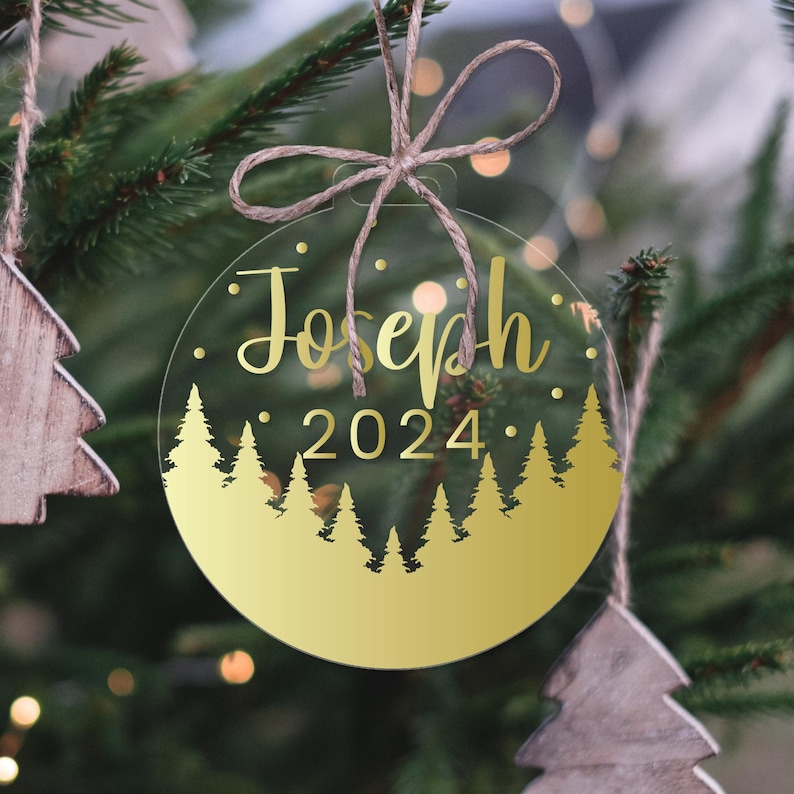 Może przedstawiać: Okrągła, przezroczysta ozdoba choinkowa z wzorem z złotej folii. Ozdoba przedstawia złotą sylwetkę lasu z sosnami i tekst "Joseph 2024".