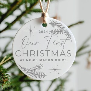 Op de afbeelding: Ronde witte kerstbal met de tekst "Our First Christmas at No. 83 Mason Drive" en het jaar 2024. De bal is versierd met gestileerde dennentakken en sterren.