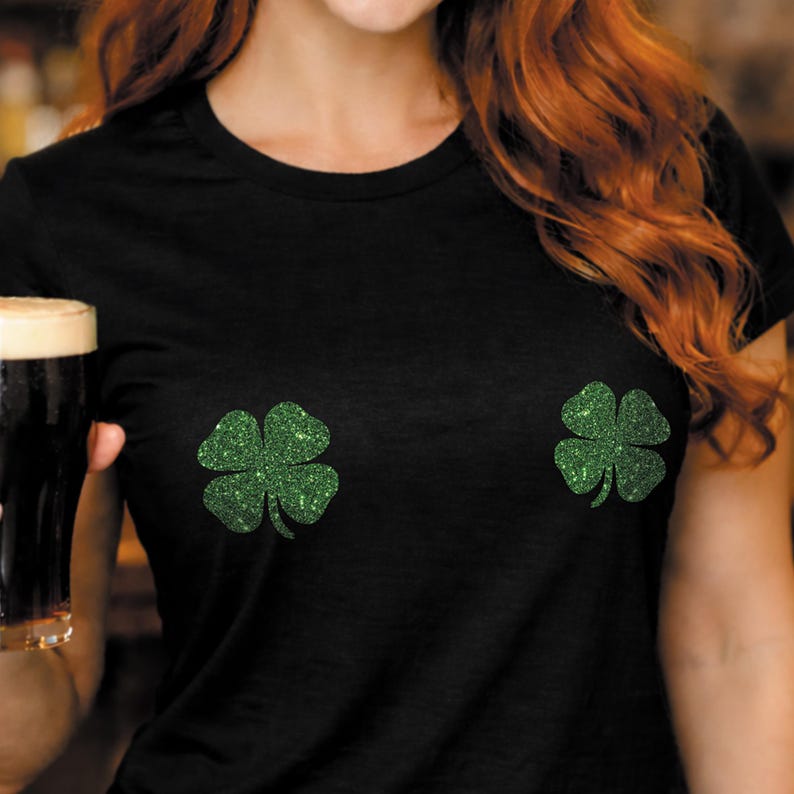May include: Black t-shirt with two green glitter shamrock designs. The shirt is a crew neck style and is made of a soft material. The shamrocks are a bright green color and are placed on the front of the shirt.