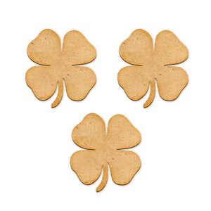 May include: Three wooden four-leaf clovers arranged on a white background. Each clover has four rounded leaves and a stem. The clovers are a light brown color and appear to be cut from wood.