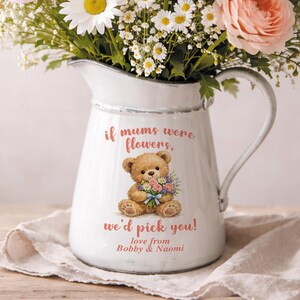 May include: White enamel pitcher with a handle, filled with a bouquet of flowers. The pitcher features a teddy bear illustration holding flowers, with the text "if mums were flowers, we'd pick you! love from Bobby & Naomi."