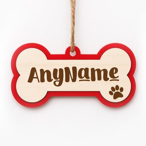 May include: A wooden dog bone-shaped ornament with a red border and a light wood center. The text "AnyName" is in brown, with a paw print on the right. A twine loop is at the top for hanging.