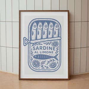 May include: A framed art print featuring a blue illustration of a sardine can. The can is labeled "SARDINE AL LIMONE" and contains sardines, lemon slices, and decorative elements. The print is in a brown frame.