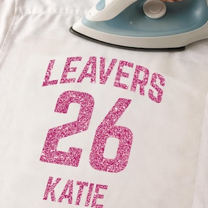 May include: A white shirt with the words "LEAVERS 26 KATIE" in sparkly pink lettering. An iron is positioned above the lettering, suggesting the application of the design to the fabric. The shirt is made of a smooth, white material.