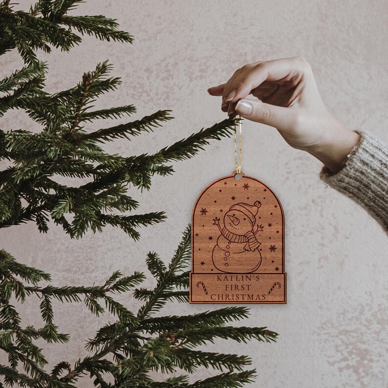 May include: A wooden Christmas bauble shaped like a snow globe with a snowman inside. The bauble is personalised with the text "Katlin's First Christmas".