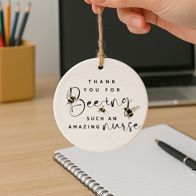 May include: A round, white ceramic ornament with a jute string for hanging. The ornament features the text "Thank You For Being Such An Amazing Nurse" with bee illustrations. The bees are black and yellow.