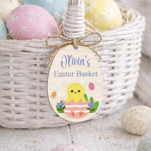 May include: A white wicker Easter basket filled with colorful decorated eggs. A wooden egg-shaped tag with the text "Olivia's Easter Basket" and a chick illustration hangs from the handle. The tag is decorated with flowers and an egg.