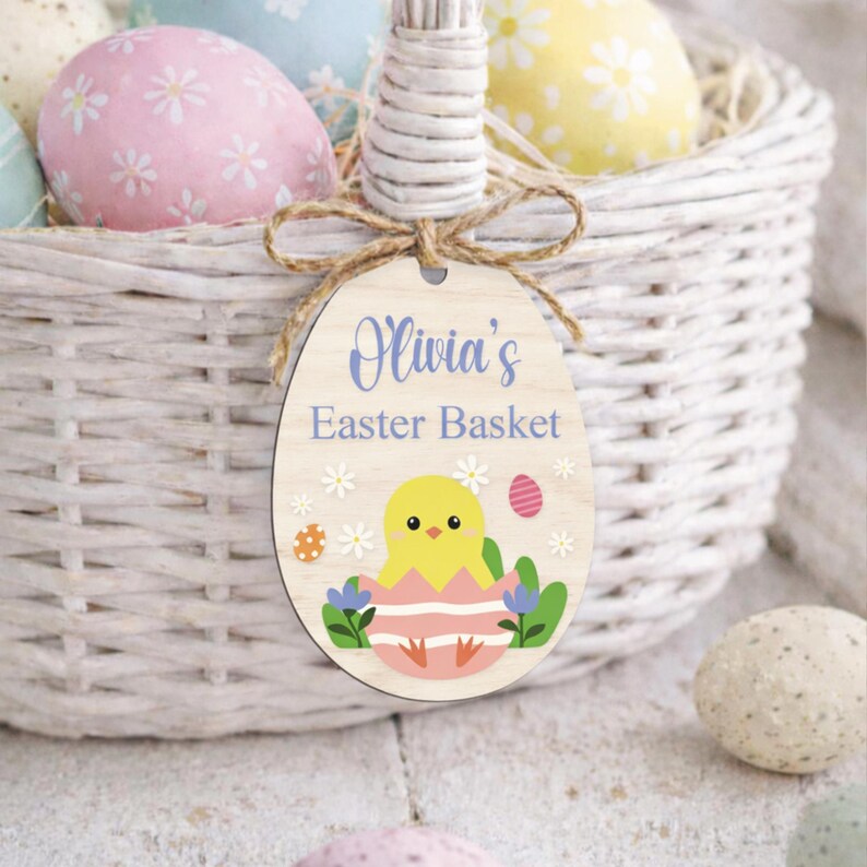 May include: A white wicker Easter basket filled with colorful decorated eggs. A wooden egg-shaped tag with the text "Olivia's Easter Basket" and a chick illustration hangs from the handle. The tag is decorated with flowers and an egg.
