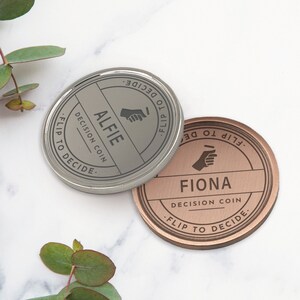 Puede incluir: Se muestran dos monedas de decisi&oacute;n, una plateada y otra cobriza. La moneda plateada est&aacute; grabada con "ALFIE" y la cobriza con "FIONA". Ambas monedas tienen el texto "DECISION COIN" y "FLIP TO DECIDE" en los bordes.