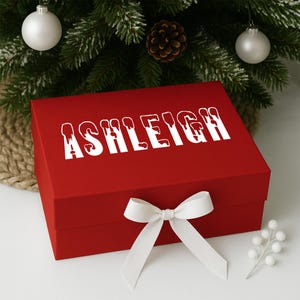 以下が含まれることがあります： 赤いギフトボックス。 蓋には白い文字で「ASHLEIGH」と書かれています。 白いリボンと蝶結びで結ばれています。 背景には、オーナメントと松ぼっくりが付いたクリスマスツリーがあります。