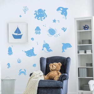 Op de afbeelding: Blauwe wanddecoraties met zeedieren, waaronder een narwal, een octopus, een schildpad, een kwal, een dolfijn, een rog, een krab, een zeepaardje en zeewier. De stickers zijn op een witte muur met een teddybeer die in een blauwe fauteuil zit.