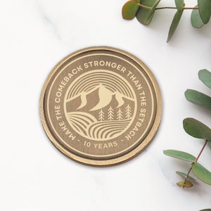 May include: A gold-colored round coin with the text "Make the comeback stronger than the setback - 10 years" and a graphic of a mountain range with trees.