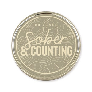 May include: A silver-colored round coin with a beige background and a white outline. The coin has a topographic design and the text "00 YEARS Sober & COUNTING" in white.