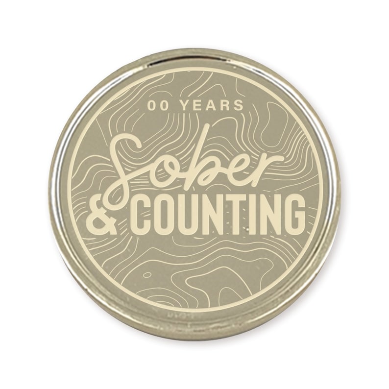 May include: A silver-colored round coin with a beige background and a white outline. The coin has a topographic design and the text "00 YEARS Sober & COUNTING" in white.