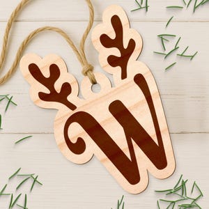 May include: Wooden Christmas ornament in the shape of a reindeer with the letter "W" in a dark brown color. The ornament is attached to a natural twine string. Green pine needles are scattered around the ornament on a white wooden surface.